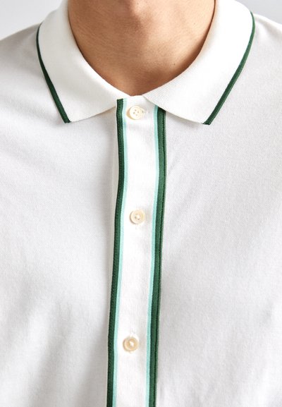 Weißes Polohemd mit grün gestreiftem Kragen und Knopfleiste, ausgestattet mit zwei cremefarbenen Knöpfen. Der Stoff hat eine strukturierte Oberfläche.