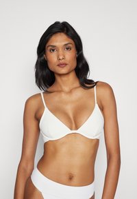 Bílý žebrovaný bikiny top s tenkými nastavitelnými ramínky, s trojúhelníkovým tvarem košíčků a hladkým, bezšvovým designem.