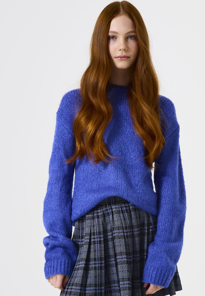 Blauer gestrickter Pullover mit rundem Ausschnitt und langen Ärmeln, kombiniert mit einem schwarz-grauen karierten Rock mit Falten.