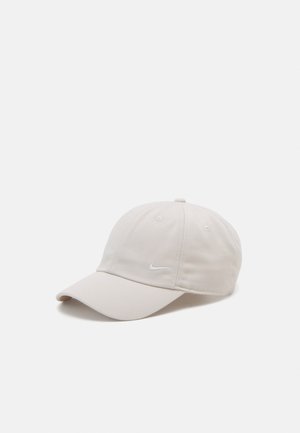 Lyse beige Nike baseballkasket med buet skygge og lille hvid swoosh-logo på frontpanelet mod ensfarvet baggrund.