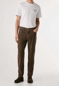 Homme portant un t-shirt blanc à manches courtes et un pantalon en velours côtelé marron avec des bottes en daim marron foncé, debout avec une main dans la poche.