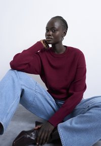 Jeune personne à la peau foncée portant un pull bordeaux et un jean bleu, assise par terre, appuyant la tête sur la main, chaussée de bottes marron foncé.