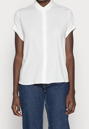 Person wearing a plain white short-sleeve collared shirt with rolled sleeves and blue denim jeans, shown from neck to hips.