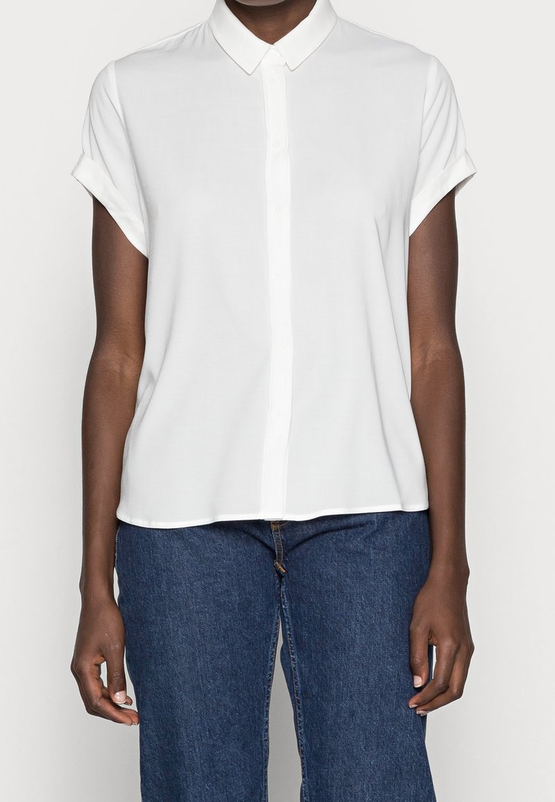 Personne portant une chemise blanche unie à manches courtes avec col, manches retroussées et jean bleu en denim, montrée du cou aux hanches.