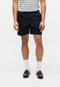 Άτομο που φοράει μαύρα σορτς, ριγέ πουκάμισο, λευκές κάλτσες και σκούρα καφέ μοκασίνια, στέκεται μπροστά σε απλό ανοιχτό χρωματικό φόντο.