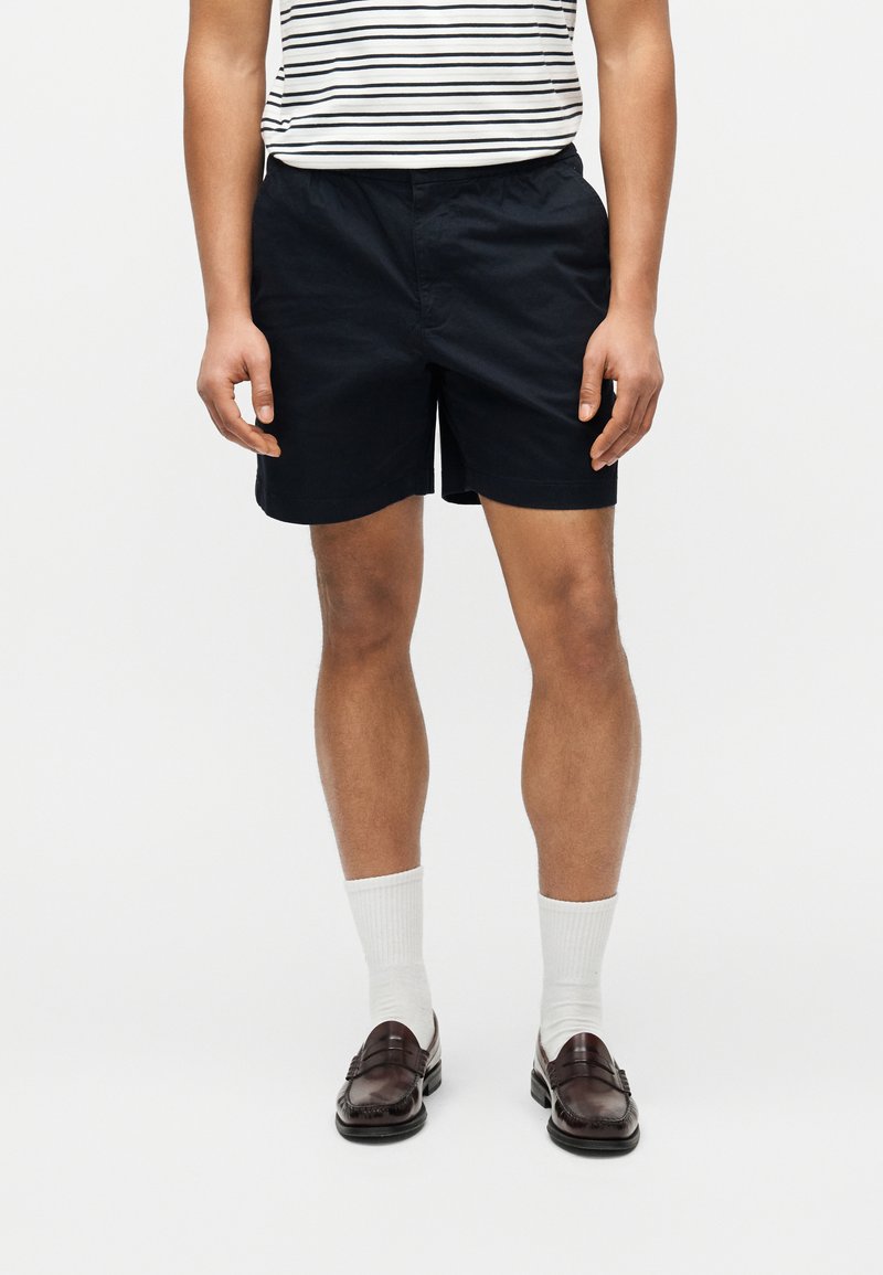 Άτομο που φοράει μαύρα σορτς, ριγέ πουκάμισο, λευκές κάλτσες και σκούρα καφέ μοκασίνια, στέκεται μπροστά σε απλό ανοιχτό χρωματικό φόντο.