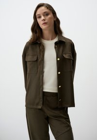 Camicia verde oliva scuro con bottoni dorati e due tasche sul petto, abbinata a un top a girocollo chiaro e pantaloni coordinati.