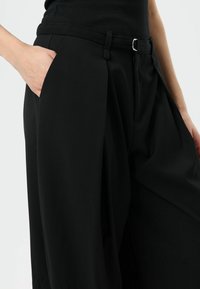 Pantalon noir taille haute avec ceinture à la taille, devant plissé et poches latérales. Le tissu semble lisse avec une forme structurée.