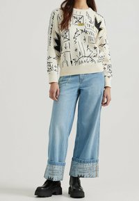 Creamfärgad tröja med svarta grafiska mönster; avslappnad passform. Ljusblå vida jeans med mönstrade muddar; kombineras med svarta kraftiga stövlar.