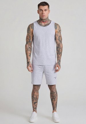 Mężczyzna z tatuażami na całym ciele, w jasnoszarej koszulce na ramiączkach i szortach oraz białych tenisówkach, stojący na jednolitym tle.