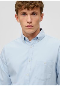 Camicia azzurro chiaro con colletto appuntito, taschino sul petto, e texture liscia, caratterizzata da cinque bottoni sulla parte anteriore.