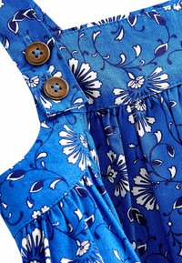 Sininen kukkakuosisekangas, jossa on valkoisia kukkakuviota, esittelee poimutetun muotoilun ja ruskeat nappikoristeet säädettävillä olkaimilla.