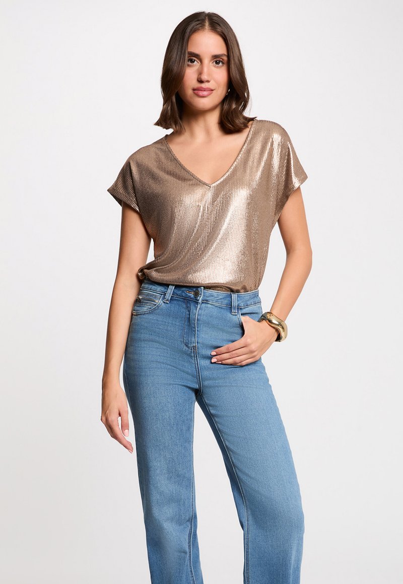 Top corto de bronce con cuello en V, que presenta una textura acanalada. Combinado con jeans de talle alto en azul claro. Pulsera dorada en la muñeca.