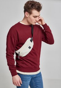 Rød hettegenser, hvit teksturert crossover-veske med glidelås og svarte detaljer, båret over en beige skjorte og blå jeans.