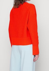 Dziany pomarańczowy sweter o luźnym kroju, z prążkowaną teksturą i szerokimi rękawami, zestawiony z jasnoniebieskimi spodniami. Widok z tyłu pokazuje szczegóły dekoltu.