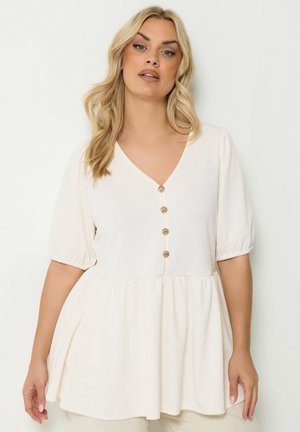 Blonde woman wearing a loose white blouse with puff sleeves and decorative shell-shaped buttons, standing against a plain background.