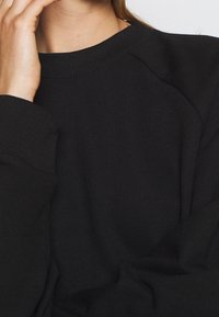 Sweat-shirt noir en coton avec un col rond côtelé et des manches raglan. Texture lisse avec un minimum de détails et une coupe décontractée.