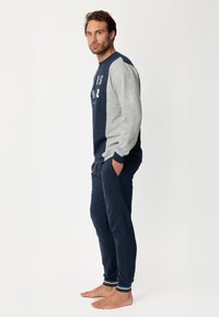 Sudadera de manga larga en azul marino y gris con texto gráfico; combinada con pantalones de chándal en azul marino con puños a rayas en los tobillos. El modelo está descalzo.