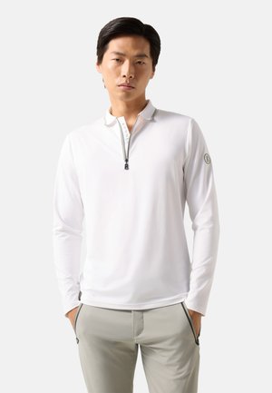 Mężczyzna ubrany w białą, długorękawową koszulę na zamek z logo na rękawie oraz jasnoszare spodnie, stojący z rękami w kieszeniach.