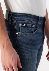 Los jeans de denim azul oscuro cuentan con un diseño ajustado, cinco bolsillos, herrajes plateados y una etiqueta con el logo negro de cK en el bolsillo delantero.