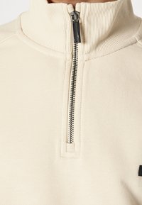 Sudadera crema con cremallera, cuello acanalado, cremallera de metal y detalle de logo negro en el frente. Hecha de tela suave y texturizada.