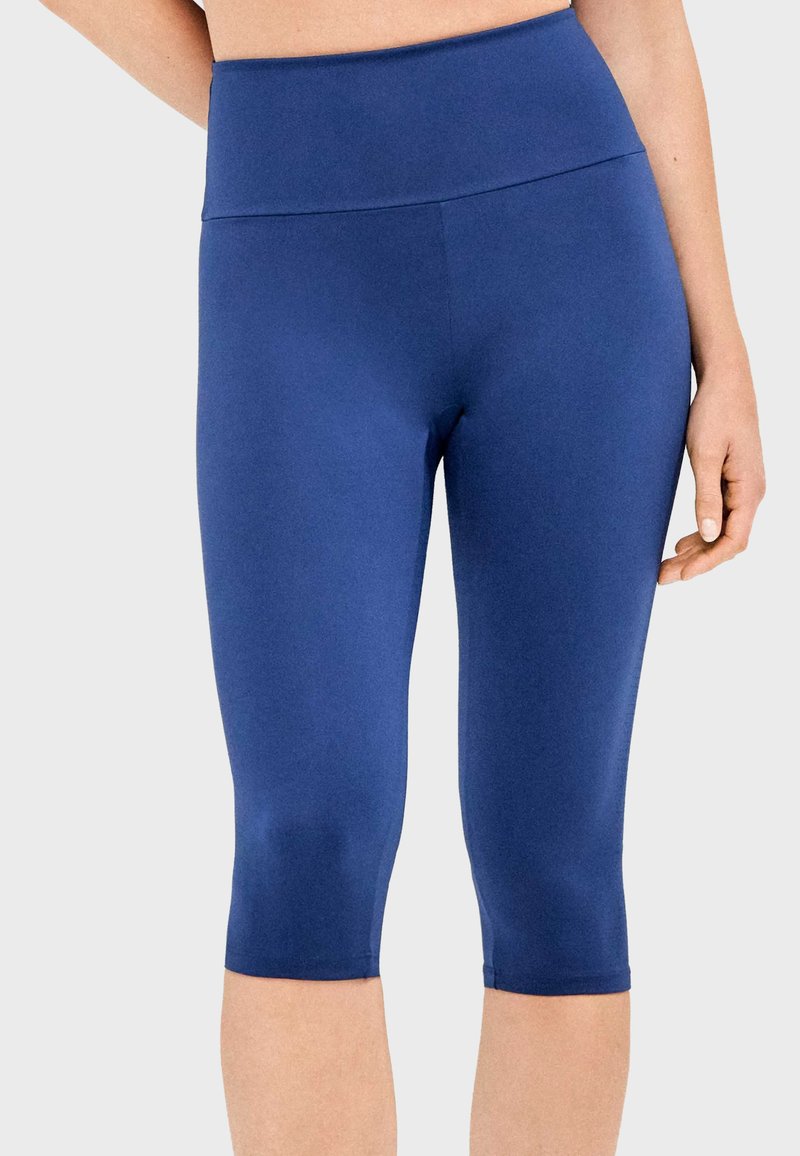 Femme portant des leggings ajustés bleu uni, taille haute, arrivant au genou, sur un fond clair uni.