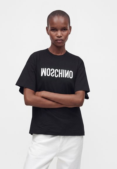 Άτομο που φοράει μαύρο μπλουζάκι Moschino με λευκό κείμενο, στέκεται με σταυρωμένα τα χέρια και φοράει άσπρο παντελόνι σε απλό φόντο.