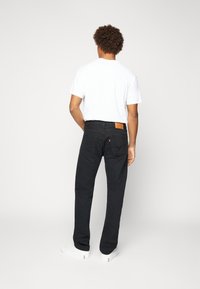 Jeans de mezclilla negros con corte recto, que presentan un parche de cuero marrón en la cintura, bolsillos traseros y se llevan con una camiseta blanca y zapatillas blancas.