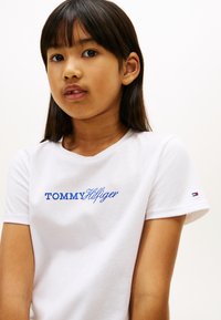 Λευκό βαμβακερό T-shirt με μπλε κεντημένο λογότυπο «TOMMY HILFIGER» στο εμπρόσθιο μέρος και μικρή ένδειξη λογότυπου στο μανίκι. Απλός σχεδιασμός.