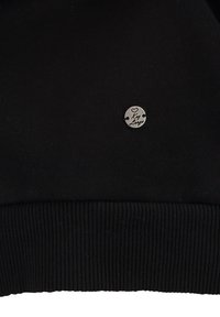 Sudadera negra con ribete en el dobladillo, textura suave y un pequeño logo redondo de plata con grabado. Diseño simple y funcional.