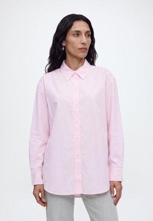 Femme aux longs cheveux noirs portant une chemise boutonnée à rayures verticales rose clair et blanche et un pantalon gris clair, debout devant un fond uni.
