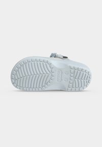 Semelle de chaussure sabot blanche à enfiler avec motif de bande de roulement texturé et sangle avec bouton noir, présentée sur un fond clair uni.