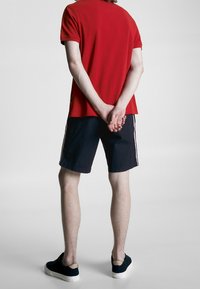 Czerwona koszulka polo z krótkim rękawem, w połączeniu z ciemnymi szortami z białymi paskami po bokach. Czarnie buty wsuwane dopełniają strój.