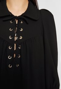 Blouse noire avec grand col et laçage devant avec des œillets argentés et des cordons noirs noués en nœud au niveau du cou.