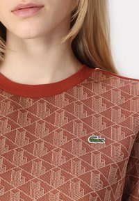Hnědý svetr se vzorem s geometrickým designem a malým zeleným logem Lacoste na levé části hrudi. Kulatý červený límec.