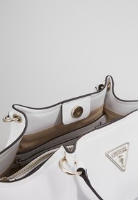 Sac à main blanc au design structuré. Présente une fermeture éclair dorée, une plaque de logo et une fermeture à pression. L'intérieur est équipé d'une doublure beige et de plusieurs poches.