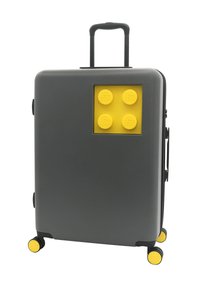 Gray hard-shell suitcase with a yellow LEGO brick design on the front, four yellow wheels, and a retractable handle for easy transport.