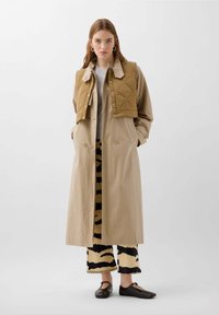 Un trench coat lung bej peste un pulover gri deschis, având un vestă căptușită maro și pantaloni cu un model în nuanțe de negru și bej; pantofi flat maro.