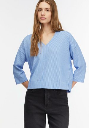 Young woman with long brown hair wears a light blue ribbed V-neck sweater and black high-waisted pants, standing with hands behind back.