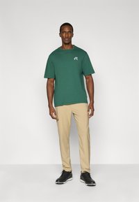 Πράσινο βαμβακερό t-shirt με στρογγυλή λαιμόκοψη και κοντά μανίκια. Μπεζ παντελόνια. Μαύρα παπούτσια. Το μοντέλο στέκεται μπροστά σε απλό γκρι φόντο.