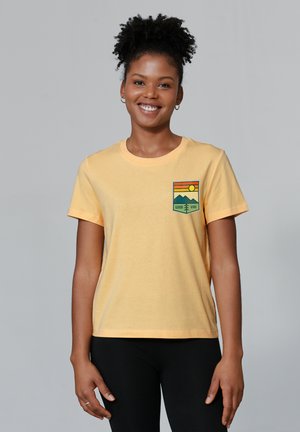 Geel katoenen t-shirt met korte mouwen, met een zakafbeelding van bergen en een zon met de tekst "GOOD VIBE" eronder.