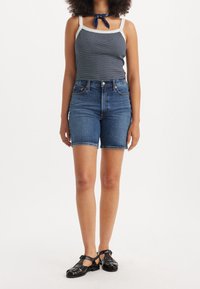 Modré denimové šortky s ohrnutými lemy, nošeny s námořnickým pruhem tank topem a černými sandály na pásky. Žádné výrazné vzory nebo kovy nejsou přítomny.