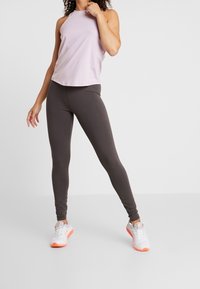 Světle fialový top bez rukávů v kombinaci s tmavě hnědými legínami, doplněný bílými teniskami s oranžovými akcenty. Hladké a elastické tkaniny.