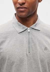 Γκρι πόλο μπλούζα με φερμουάρ, με διπλωμένο γιακά και λεπτή λεπτομέρεια λογοτύπου στο στήθος. Κατασκευασμένη από μαλακό, υφασμάτινο ύφασμα με υφή. Ασημί διακοσμητικό φερμουάρ.