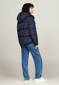 Marine pufferjack met capuchon, voorzien van gewatteerde secties en elastische manchetten, gecombineerd met lichtblauwe wijde jeans en witte sneakers.