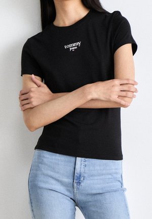 Nainen, joka pukeutunut mustaan lyhythihaiseen Tommy Jeans -t-paitaan ja vaaleansinisiin korkeavyötäröisiin farkkuihin, kädet ristiin rinnan päällä.