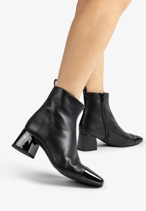 Zwarte leren enkellaarzen met blokhakken en glanzende lakneus, gedragen op blote benen, getoond tegen een witte achtergrond.