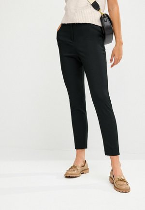 Femme portant un pantalon noir slim, des mocassins beiges avec un détail de chaîne dorée, un top en maille crème et un sac bandoulière noir avec une large bandoulière.