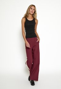 Black tank top paired with burgundy wide-leg trousers featuring side accents and a smooth texture. Model stands with hands in pockets.