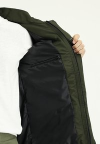 Olijfgroene jas, ritssluiting aan de voorkant, zwarte voering, met een klein ritsvakje aan de binnenkant. Zachte stof met een gladde textuur.